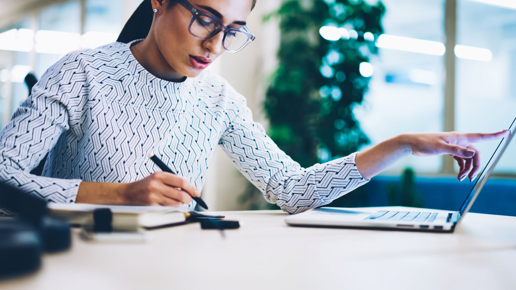 A focused woman with glasses using a laptop and taking notes at a modern office desk.